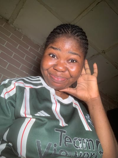 A young woman with braided hair smiles. She has a nose piercing and wears a green and white striped shirt with the Adidas logo. Her hand is positioned near her cheek. A brick wall appears in the background alongside a dark, unlit space. The image is slightly rotated, giving an unconventional perspective.