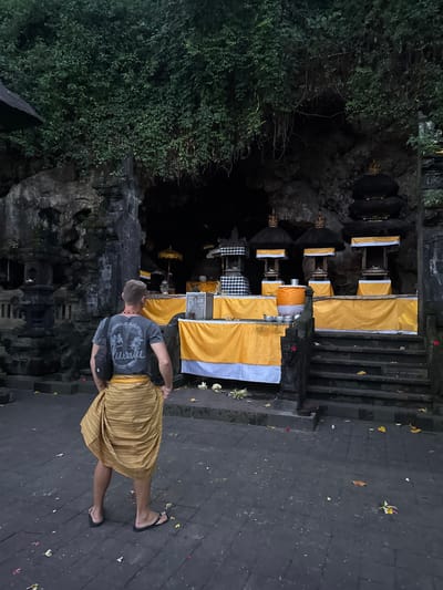 In the city of Dawan, Indonesia, a man stands facing a religious shrine in front of a cave entrance. He wears a grey t-shirt with a design, a yellow sarong wrapped around his waist, and flip-flops. A black crossbody bag is slung over his shoulder. The shrine is decorated with yellow fabric and features several tiered structures topped with dark, pointed roofs. Umbrellas with yellow fabric are also visible. The ground is paved with dark stones, and fallen leaves and flower petals are scattered across the area. The background shows a rocky cave entrance covered in lush green foliage. The lighting suggests it is either dusk or a dimly lit indoor space.
