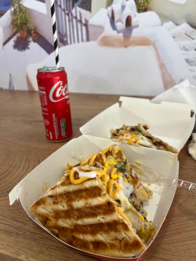 The media file shows a close-up of a meal consisting of grilled sandwiches and a Coca-Cola can with a straw. The sandwiches appear to be filled with meat, cheese, and sauces, served in white paper containers. A clear plastic fork rests in one of the containers. The Coca-Cola can is red with the iconic white logo and has a black and white striped straw inserted. The meal is placed on a wooden surface, suggesting an indoor setting. In the background, there are blurry images on white paper, possibly decorative elements or printed menus, and a glimpse of a person's hand. The lighting suggests it is daytime. The visible text on the Coca-Cola can reads "Coca-Cola" and some text in what appears to be Bulgarian, including "Вкусен" (vkusen - delicious) and "напитка" (napitka - drink). The scene appears to be casual, possibly a meal break or a snack. The location context provided is Byala Slatina, Bulgaria.