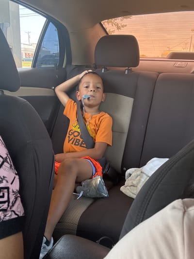A young boy is lying down in the back seat of a car, with his head resting on his arm and his body angled towards the viewer. He is wearing an orange t-shirt that says "Good Vibes" in white letters, and blue and orange shorts. He has a nasal cannula in place, with tubing running from his nose to a device attached to his shirt. He is also wearing grey sneakers with white laces. The car's interior is visible, including the dark grey seats and a seatbelt. Outside the car, through the window, a bright, sunny day is suggested by the strong light and clear sky. The scene appears to be in Balch Springs, United States of America. The boy's expression is neutral, and the overall mood of the image is calm.
