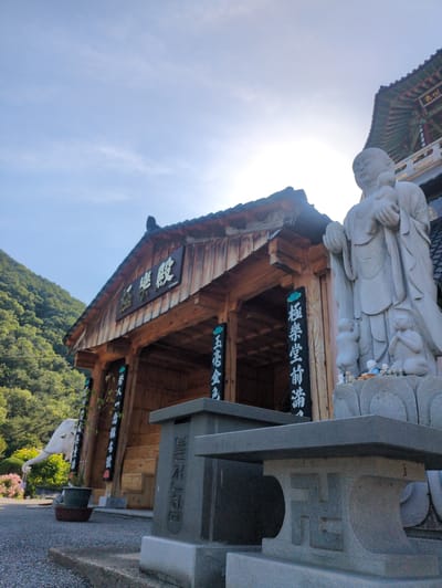 The image displays a Buddhist temple complex located in Godang-ri, South Korea, under a bright, partially cloudy blue sky. A dense, verdant mountain range covered with trees forms the background.

In the immediate foreground on the right, a large, light-colored stone statue of a standing Buddha figure holds a smaller infant figure. Two smaller, adolescent-like figures are seated at the base of the main statue; one is prostrate, and the other kneels with hands together. Multiple miniature, doll-like figures are arranged on the base platform in front of the larger statue. To the left of this statue, a gray stone table-like structure features a carved Buddhist swastika symbol on its front support. Adjacent to this, a taller, rectangular gray stone pillar displays vertically carved Korean characters.

In the midground, a traditional Korean wooden building with an open porch and a dark, multi-tiered tiled roof is prominent. A rectangular dark sign with three large Korean characters, identified as '極樂殿' (Geungnakjeon, meaning "Hall of Supreme Bliss"), is mounted above the building's entrance. Two vertical black banners with white Korean characters hang from the facade; the banner closer to the Buddha statue reads '極樂堂前滿' (Geungnakdang Jeonman), and a partially visible banner further left reads '五臺金山' (Odae Geumsan). A potted plant containing a sapling is placed on the ground in front of the building. Further left, a large white statue of an elephant with an uplifted trunk is partially visible behind green foliage and pink flowering bushes.

The ground surface in the foreground and midground is composed of gravel. Bright sunlight is evident, creating a prominent lens flare in the upper right quadrant of the image and causing some overexposure in the sky. No active human subjects are visible within the frame.