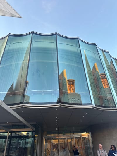 A modern building with a distinctive curved glass facade reflects surrounding architecture. The glass panels are stacked horizontally, creating a wave-like pattern. The reflection captures a tower with a rounded top. The building's entrance features glass doors, and the words "ANNA & JOHN J. SIE WELCOME CENTER" are visible on the wall above. People are near the entrance, adding scale. A white, pointed structure appears to the left of the building, contrasting with the blue sky.