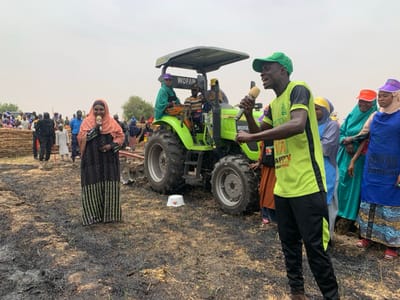 The image shows a group of people standing in a field, the ground covered in burnt stubble, in Ringimawa, Nigeria. A green tractor with the word "WOFAN" printed on it is parked nearby. A woman in a dark dress with a pink scarf is standing to the left of the center, speaking into a microphone. A man in a green shirt with the word "HAPPY" on it is speaking into a microphone, standing to the right of the center. Other people, including children, are gathered around. The sky is overcast and the day appears to be cloudy.