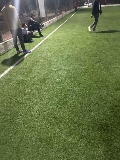 An artificial turf soccer field is depicted with a white sideline marker. Several people stand along the sideline next to a chain-link fence. The field itself is a vibrant green and appears well-maintained. Some of the people are wearing athletic shoes, suggesting they may be involved in playing or training on the field. The lighting suggests it is either dusk or an artificially lit field.
