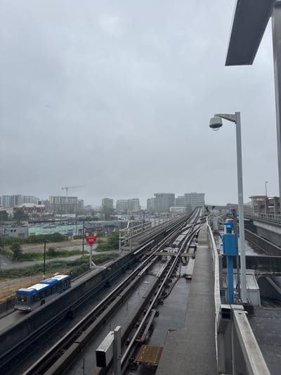 The image depicts an elevated train track winding through a city with overcast skies. A blue and white train is visible on the tracks, stopped or moving slowly. The scene appears to be in Richmond, Canada, given the context. The weather is gloomy and rainy, with visible water droplets on the train and tracks.

Several objects are visible: a security camera mounted on a pole, a blue signal box, and various structures and buildings in the background, including high-rise buildings and construction sites with a crane. There is a red hexagonal sign with white text that reads "SWITCH 325". The train has "302" displayed on its back. The overall atmosphere is one of a typical urban environment on a dreary, wet day. There are no people clearly visible in the image. The image is taken from a high vantage point, looking down onto the train tracks and the city.