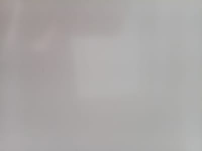 This image is a close-up, slightly out-of-focus shot of a light gray, textured surface. The surface appears to be indoors, possibly a wall or a piece of fabric. There are subtle variations in tone and slight shadows, suggesting ambient lighting. No discernible subjects, activities, or text are present. The scene is abstract and devoid of specific context.