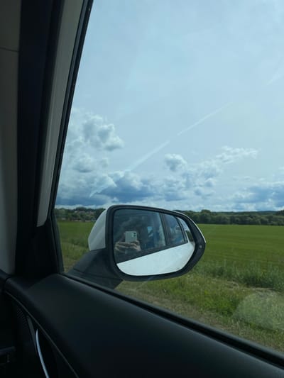 The image depicts a view from inside a car, looking out the passenger side window. The primary subject is the side-view mirror, which reflects a person holding a smartphone. The mirror is framed by the car's interior, featuring black plastic trim. Outside the car, a rural landscape unfolds under a cloudy sky. Rolling green fields stretch towards a line of trees in the distance. The reflection in the mirror suggests it is daytime, with bright but diffused light, indicating a partly cloudy or overcast day. The overall scene conveys a sense of travel through a natural, open environment. No visible text is present in the image.