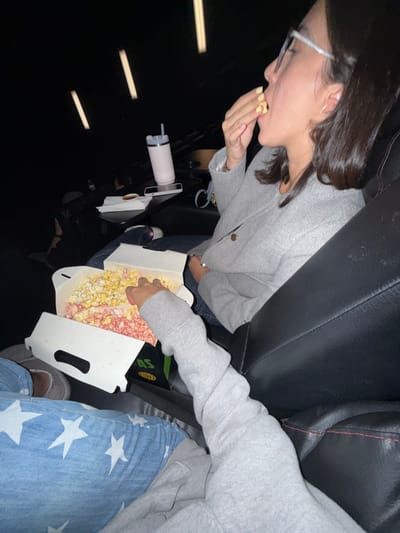 The image depicts an indoor scene within a cinema located in La Purísima, Mexico. Two individuals are seated in dark, upholstered cinema seats.
To the right, an adult female is shown in profile, from the chest up, with dark hair and eyeglasses. She wears a grey long-sleeved top or jacket. Her right hand, featuring light yellow nail polish, is raised to her mouth, indicative of eating. A large, white cardboard popcorn box with an open lid is positioned between the two individuals, containing multi-colored popcorn, primarily yellow/white and pink/red varieties. A green and black logo is partially visible on the side of the box.
To the left, a second individual is partially visible. Their left leg is clad in blue pants with white star patterns, and their left arm, covered by a grey long-sleeved garment, is extended into the popcorn box.
On a small table or armrest to the left of the female, a tall, light-colored insulated cup with a dark lid and straw is present. Beside it are a white smartphone or similar device, a small dark dish, and a white napkin or wrapper. The background is dimly lit, revealing out-of-focus rows of cinema seats and several vertical light fixtures characteristic of theater lighting. The overall environment is dark, suggesting a film screening is in progress.