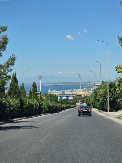 The image depicts a view from a road descending towards the city of Thessaloniki, Greece. In the foreground, a black sedan is driving away from the camera on the asphalt road. The road is flanked by lush green trees and flowering bushes on both sides. In the middle ground, the city of Thessaloniki is visible along the coastline of the Aegean Sea, with various buildings and structures, including a stadium with floodlights, a port with ships, and a prominent tower. The background features a hazy mountain range and a clear blue sky with a few scattered clouds. The overall scene suggests a sunny day in an urban coastal environment.