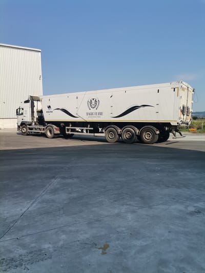 A white semi-trailer truck with the brand name "MAGIC FLAME" printed on its side is parked on a concrete surface. The truck has a long, white trailer with several wheels. Next to the trailer, a white industrial building with a corrugated metal exterior is visible. The sky above is clear and blue. The scene appears to be an outdoor industrial or commercial area, possibly a logistics hub, in Slanchevo, Bulgaria.