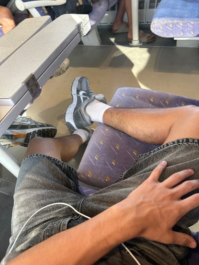 This is a first-person perspective image taken from a seat inside what appears to be a train car, possibly in the city of Rivesaltes, France, given the context. The foreground features the legs of a person wearing dark denim shorts and white socks with athletic shoes. A white wire, possibly for headphones or a charger, is draped across the shorts. To the left, a grey plastic tray table is visible, attached to the seat in front. In the middle ground, the legs of another person are seen, wearing sandals. The seats in the background are upholstered in a purple fabric with a subtle pattern of yellow and white lines. The floor of the train car appears to be grey. The lighting suggests it is daytime, with sunlight casting shadows.