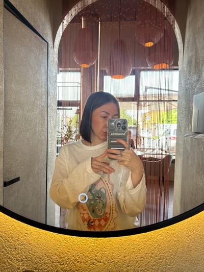 The image captures a mirror selfie taken by an adult female within a commercial establishment in Усады, Russia, likely a restaurant, cafe, or its associated restroom. The woman is positioned centrally, visible from the chest up, holding a dark gray smartphone vertically with both hands to capture her reflection. She has straight, dark, mid-length hair parted on her left side (viewer's right). Her facial features are partially obscured by the phone, but her right eye, nose, and mouth are discernible as she appears to be looking at the phone screen. She wears a white, long-sleeved top with a translucent quality, featuring a colorful graphic design on the lower left, depicting abstract patterns, a star, and checkered elements. On her left hand (viewer's right), she wears a prominent silver-toned ring with a central stone on her index finger, multiple stacked band rings on her middle finger, and a thin band ring on her ring finger. Her right hand (viewer's left) displays a single thin band ring on the ring finger. The smartphone, likely an iPhone, features a triple camera array on its rear and is housed in a textured, ribbed protective case. The mirror itself is oval or rounded-rectangular with a thick black frame along its bottom edge. This lower section of the mirror emits a warm, textured golden light. A small, circular, illuminated blue-white touch control or light source is reflected on the mirror's surface near the woman's lower left arm. The background, visible in the mirror's reflection, details the interior and exterior environment. The walls appear textured: a light brown/beige on the left with a distinct dark vertical line, possibly a door frame, and a black horizontal handle, suggesting a door. On the right, the wall is a lighter gray textured surface, featuring a wall-mounted silver-colored soap or sanitizer dispenser. An arched opening is visible in the upper right section, leading to another internal area. Directly behind the woman, and extending across the upper portion of the reflection, is a decorative bead curtain composed of numerous thin, reddish-brown strings with small, teardrop-shaped beads. Through and above this curtain, several large, spherical or oblong pendant lights are visible, emitting a warm orange glow from their bases. Beyond the bead curtain, a window or open area reveals an outdoor scene under bright daylight. Green foliage, an outdoor seating area with tables and chairs, and a partially visible white vehicle (possibly a