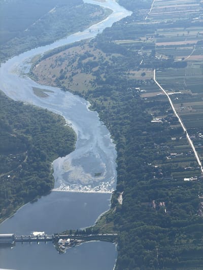 This aerial photograph depicts a wide river with a dam and a hydroelectric power station located on its southern bank. The river curves from the northwest to the southeast. On the east bank of the river, agricultural fields are visible, interspersed with forested areas and a road. Houses and other buildings are scattered throughout the landscape. The dam features a road crossing over it, with vehicles present. The hydroelectric power station is a large, concrete structure. The area appears to be rural or semi-rural, with evidence of human settlement and land use. The lighting suggests it is daytime, with some atmospheric haze. The location is identified as Ziąbki, Poland.