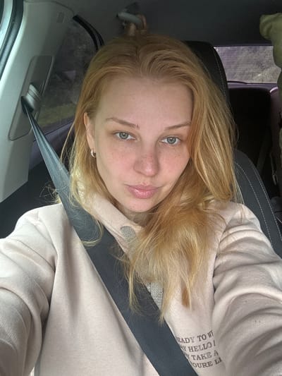A young woman with blonde hair and freckles is sitting in the driver's seat of a car, wearing a light pink hoodie and a seatbelt. Her eyes are green, and she has a neutral expression. The interior of the car is visible, including the dashboard, steering wheel, and a seatbelt. Outside the car, trees and foliage can be seen through the window, suggesting a rural or suburban setting, possibly on the outskirts of Novoe, Russia, given the context. The lighting is natural and appears to be daylight. The image is a selfie, taken from a slightly high angle.