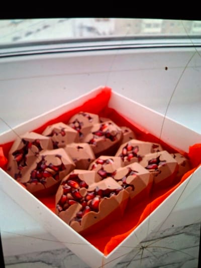 A white box filled with faceted chocolate candies, each embedded with pomegranate seeds, is positioned on a windowsill. The box is lined with bright red fabric. Outside the window, a blurred cityscape of buildings is visible. The scene appears to be indoors, likely a domestic setting, in the city of Medvezhyi Ozyora, Russia.