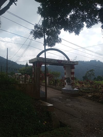 The scene is depicted in the city of Niz Lakuk, India. The image presents an outdoor, static view of an arched gateway spanning a dirt path under an overcast sky. The gateway consists of two substantial pillars, each painted with horizontal red and white stripes, supporting a plain gray concrete arch. Black graffiti marks are visible on the lower portion of the right pillar. Incomplete text reading "BUJOL CIJREUM KALER" is inscribed on the crossbeam above the pillars. The dirt path, showing signs of erosion and unevenness, passes directly through the arch and recedes into the background. To the immediate left, a dense growth of green foliage covers a raised earth embankment. Utility poles and multiple overhead power lines crisscross the upper portion of the frame against a cloudy sky. Beyond the gateway, the landscape features scattered vegetation, including plants with reddish leaves, and distant, heavily forested hills. A metal fence is partially visible to the left of the gateway, behind which a small structure with a reddish roof is discernible. No individuals are present in the image.
