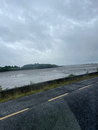 The image shows a roadside scene in Tullycullion, Ireland, on a cloudy day, likely during or after rainfall. The perspective is from a vehicle, looking diagonally across a wet road towards a large, flat body of water. The road is dark asphalt with visible yellow lane markings, suggesting it's a public thoroughfare. The water appears to be a lake or marshy area, reflecting the overcast sky, and is bordered by a strip of green grass and vegetation. In the background, a lush, tree-covered hillside rises, characteristic of the Irish landscape. The sky is filled with grey, textured clouds, indicating a dull and possibly humid atmosphere. There are no people or specific objects to identify, and the overall impression is one of a natural, somewhat desolate, and wet environment. The time of day is indeterminate due to the overcast conditions, but the light suggests daytime.