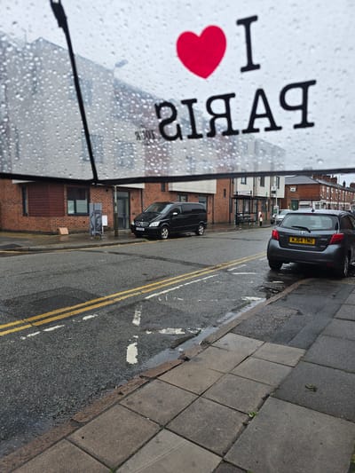 A clear umbrella with water droplets on it is held in the foreground, obscuring parts of a street scene. The umbrella has "I ❤️ PARIS" written on it, with "PARIS" appearing in reverse. The street is wet and appears to be in a residential or commercial area, with overcast skies.
On the street, there is a dark-colored Mercedes-Benz van parked on the left, with license plate "T32 XGX". To the right of the van, across the street, a dark grey Toyota Yaris hatchback is parked, with license plate "MJ64 TMO". The Yaris is facing away from the viewer and its rear is visible.
The buildings in the background are brick structures with windows and a bus stop shelter. The road has double yellow lines painted on it and some puddles reflecting the light. The sidewalks are made of concrete paving slabs.
The scene is likely a typical urban street in Leicester, United Kingdom, given the style of the buildings and the car license plates. The wet conditions suggest it might be raining or has recently rained. No people are visible in the image.