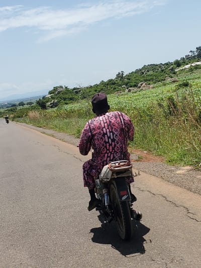 A person wearing a dark cap and a pink and black patterned outfit rides a motorcycle on an asphalt road. The motorcycle has a passenger seat with some bags attached. The road is bordered by tall green grass and a field of green crops on the right. In the background, there is a rocky, vegetated hillside. In the distance, mountains are visible under a partly cloudy blue sky. Another person on a motorcycle is further down the road to the left. The scene appears to be in a rural or semi-rural area, consistent with locations around Pankshin, Nigeria.