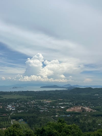 An elevated, wide-angle, vertical shot captures a panoramic view of Baan Thurian, Thailand, under a dynamic sky filled with large, white cumulus clouds. In the foreground, the tops of lush green trees are visible. The midground reveals a sprawling landscape dominated by a small city nestled amongst dense, green vegetation. Buildings with a variety of colored roofs, roads, and what appear to be agricultural fields are scattered throughout. A prominent, lighter-colored hill or quarry site is visible in the middle ground, featuring exposed rock. Beyond the city and the varied terrain, a body of water, likely the sea, stretches to the horizon, with islands and distant, hazy mountains forming the backdrop. The sky is a mixture of blue and white, with the large cloud formation drawing significant attention due to its size and dramatic appearance. Two small, dark specks, possibly birds, are present in the upper portion of the sky. The overall impression is of a vast, natural landscape punctuated by human settlement.