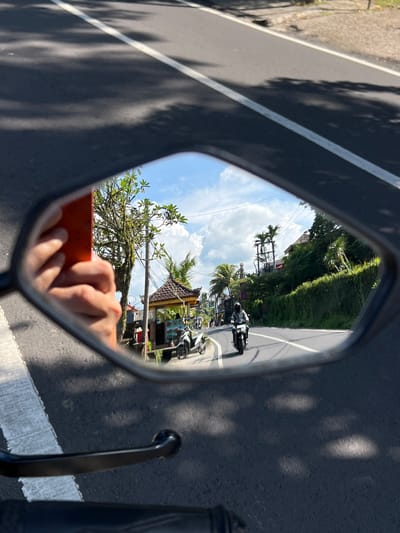 The image shows a view reflected in a motorcycle side mirror. The mirror's reflection includes a road in Petulu, Indonesia, lined with tropical vegetation and buildings. Two motorcycles are visible on the road behind the photographer. The photographer's hand, holding an orange-cased device (likely a phone), is partially visible in the lower portion of the mirror's reflection. The immediate foreground of the image shows the motorcycle's handlebars, mirror frame, and the road upon which the motorcycle is traveling. The scene is brightly lit.