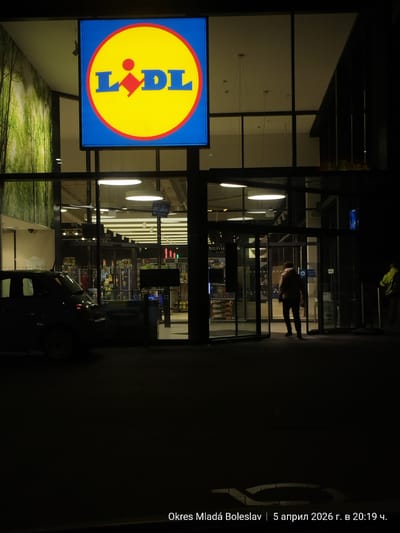 Here is a forensic description of the image:

The image captures the entrance of a Lidl store at night in the Okres Mladá Boleslav region in Czechia, dated April 5, 2026, at 20:19. The prominent Lidl logo, consisting of the word "LIDL" in blue lettering within a yellow circle outlined in red against a blue square, is positioned above the entrance. The store features large glass windows and doors, reflecting the interior lights. A person wearing a jacket and dark pants is seen entering the store. A parked black car and a green recycling bin can be seen. The setting is dark, with minimal ambient light apart from the store's illumination. There is also a security guard in a yellow jacket near the entrance.
