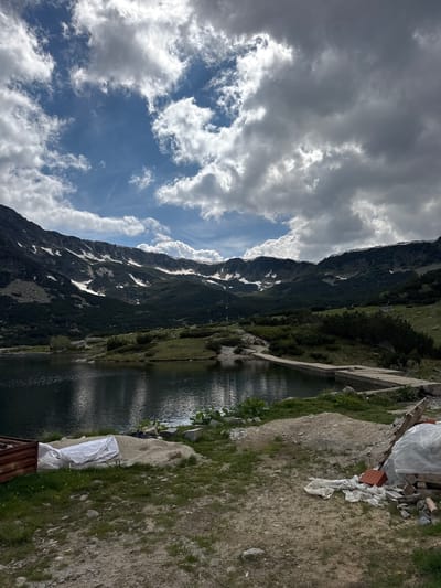 The image depicts a scenic mountain lake landscape in Avramovo, Bulgaria. The foreground shows a gravelly area with some construction debris, including a white tarp, a rusty metal structure, and piles of sand and bricks. A dark, still lake occupies the middle ground, reflecting the sky and surrounding mountains. The far background is dominated by a majestic mountain range, with dark green pine forests covering its lower slopes and patches of snow visible on the higher elevations. The sky is a dramatic mix of bright blue and heavy, white and grey clouds, suggesting dynamic weather. The scene is natural and serene, with a touch of human presence in the form of the construction materials in the foreground.