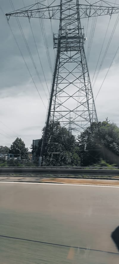 The image captures a view from a moving vehicle, likely on a highway, in Endersbach, Germany. The primary subject is a tall, metal electricity pylon with numerous cables extending from it, dominating the upper portion of the frame. The pylon is a lattice structure, typical of high-voltage transmission towers.
In the middle ground, behind the pylon, is a dense line of trees and foliage, suggesting a natural or park-like area bordering the road. Partially visible behind the trees and to the left of the main pylon is a building with a cylindrical structure, possibly a silo or industrial facility. Further in the distance, another electricity pylon is faintly visible, indicating a network of power lines.
The foreground consists of a blurred representation of a road surface, characterized by distinct lane markings and a gray barrier, emphasizing the motion of the vehicle from which the photo was taken. The sky is overcast with a muted, grayish tone. The overall impression is one of industrial infrastructure within a semi-urban or peri-urban landscape in Endersbach. There are no people or animals clearly visible in the image. The focus is on the power transmission infrastructure and the surrounding environment as seen from a moving perspective.