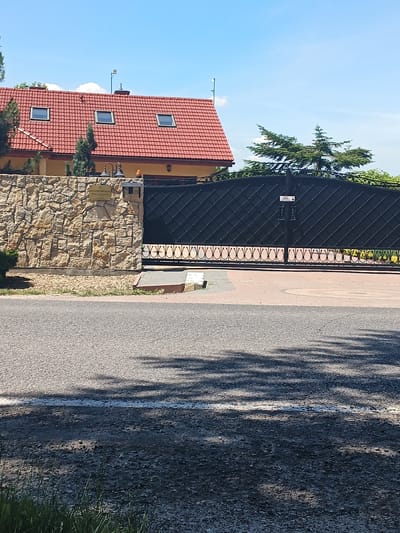 The image depicts a closed, ornate black metal gate in front of a stone wall and a red-tiled roof house. The gate features a diamond pattern and decorative scrollwork at the bottom. A small white sign with black text is affixed to the gate. Above the gate and stone wall, the house has a red tile roof with three dormer windows and is bordered by green trees. To the right of the house, a large conifer tree is visible. The sky is bright blue with a few wispy clouds, suggesting a clear and sunny day. The foreground shows a paved road with a white line, and shadows are cast across the road, indicating it is daytime. A small sign with text is mounted on the stone wall to the left of the gate. The setting appears to be a residential property in a suburban or rural area, possibly a driveway entrance to a home or a business. The overall mood is serene and tranquil. The text on the sign on the stone wall appears to read: "BANCZAROWA KOMPLAKSA Maria & Bogdan Zaborowscy".