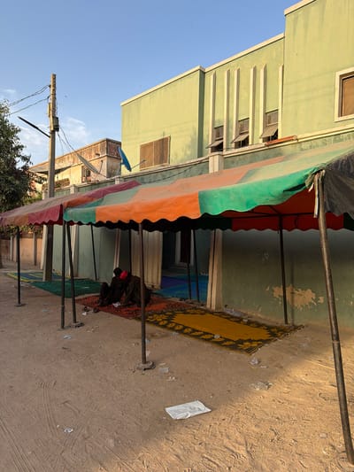 The scene depicts an outdoor area in Daurawa, Nigeria, under clear blue sky with direct sunlight. The ground is unpaved, consisting of light-colored sand and dirt, exhibiting tire tracks and scattered litter, including plastic bags and wrappers.
A series of five connected, weathered fabric awnings, featuring segments of red, orange, green, and black fabric, extend horizontally across the mid-ground. These awnings are supported by dark metal poles. Beneath the awnings, multiple patterned rugs and mats are laid out on the sandy surface, leading towards a building. The closest visible rug is predominantly red with black and yellow patterns, followed by a yellow rug with black floral patterns. Further back, blue and green mats are visible at an entryway.
Two individuals are seated on the red patterned rug under the leftmost awning. Both are dressed in dark clothing. One individual, positioned on the left, wears a red head covering. They appear to be engaged in an activity or interaction on the rug.
In the background, a two-story building with a pale green facade is prominent. Its upper section features three vertical, light-colored decorative elements and multiple windows, some of which are tilted open. The lower wall exhibits wear and peeling paint. To the left and slightly behind this structure, an older, lighter-colored building is visible, featuring a blue satellite dish on its exterior. Another blue satellite dish is mounted on the green building. A wooden utility pole, heavily laden with electrical or communication wires, stands on the far left. A small tree or bush is partially visible behind the utility pole.