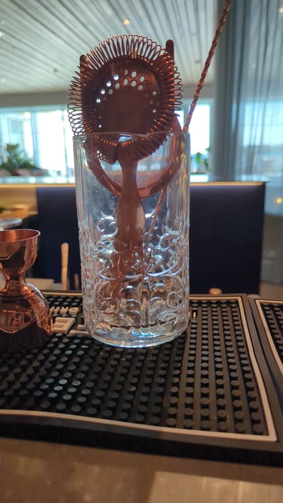 The image shows a copper Hawthorne strainer and a copper bar spoon resting inside a clear, patterned glass on a black bar mat. To the left of the glass is a copper jigger. In the background, through a window, greenery and a modern interior with wood paneling are visible. The scene appears to be set up for drink preparation, likely in a bar or restaurant. The location is identified as Larkollen, Norway, based on the information provided. The image is a medium close-up, focused on the bar tools and glassware.