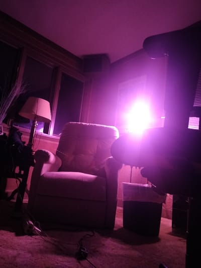 The image depicts an indoor residential room in Roswell, United States, illuminated primarily by a pervasive purple-magenta light. A strong, bright light source emanating from the lower right foreground creates significant lens flare across the scene. The room features a light-colored, upholstered recliner chair positioned centrally. To its left, a floor lamp with a beige-colored shade stands near two large, dark-appearing windows. A dark-colored side table with indeterminate objects on top is partially visible to the far left. On the right side of the recliner, a dark trash can with a white plastic liner is situated on the carpeted floor. Several dark electrical cords, including one connected to a power strip, lie on the foreground carpet. A dark rectangular speaker is mounted high on the upper-left wall. A framed object is visible on the wall between the recliner and the intense light source. Tall, slender, light-colored decorative branches are present on the far left. The flooring is carpeted, and the walls appear to be dark. No human subjects or active events are depicted.
