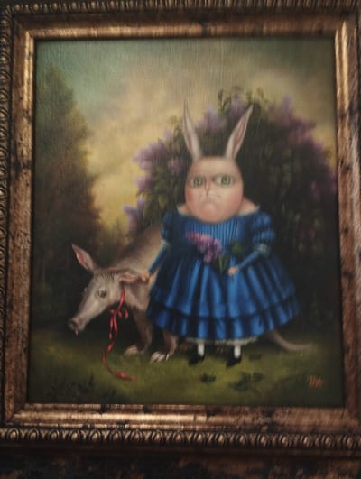 This is a framed oil painting of a surreal scene. In the foreground, a light-colored, animalistic creature with a long snout and large ears, possibly an aardvark or a piglet, stands to the left. It has a red ribbon tied around its neck. To its right, a humanoid figure with rabbit ears and a round face is depicted wearing a ruffled blue dress. This figure holds a bouquet of purple flowers and appears to have a stern expression. The background shows a lush green landscape with trees and a large cluster of purple flowers, suggesting a garden or park setting. The painting is enclosed in an ornate, gold-colored frame.
This image was taken in Pardes Hana-Karkur, Israel.