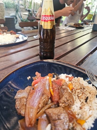 The image depicts a partially consumed plate of Lomo Saltado, a traditional Peruvian dish, consisting of stir-fried beef, onions, tomatoes, and rice on a dark blue plate. A fork rests on the plate near the rice. A brown bottle of Cusqueña beer lies horizontally on the wooden table, partially obscuring a wine glass and another plate of food in the background. In the background, two people are visible, one in a black shirt and the other in a white top. The scene appears to be set outdoors, potentially on a patio or deck with a wooden table. The location is La Molina, Peru.
