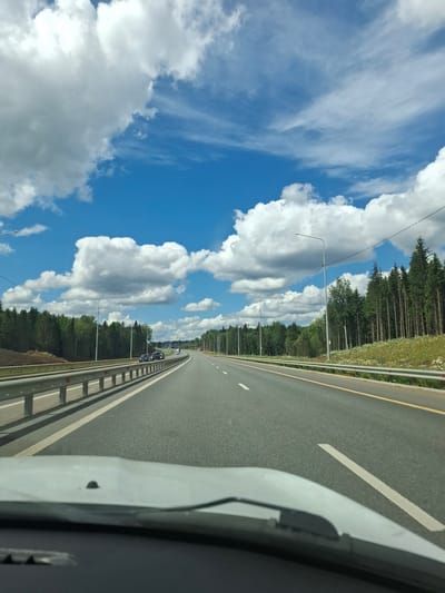 This is a POV shot from a vehicle driving on a highway. The highway has two lanes in each direction, separated by a median. Guardrails line the side of the road, and trees and a grassy embankment are visible beyond the guardrails. The sky is bright blue with scattered white clouds. There are streetlights lining the highway. A few cars are visible on the road ahead. The foreground shows the dashboard and windshield of the vehicle. The image was taken in Urazgil'dy, Russia.