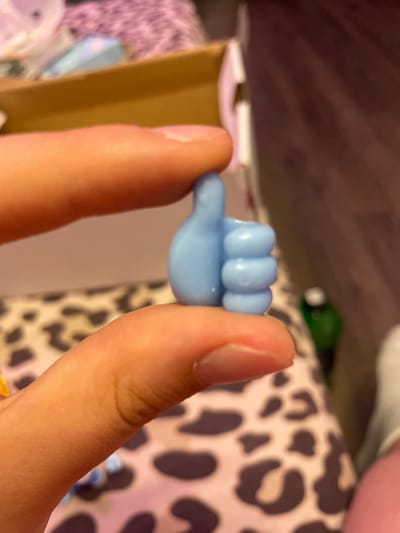 In a close-up shot, two fingers, one above and one below, hold a small, light blue, thumb-shaped object. The object has three distinct segments on the thumb, indicating it may be a miniature representation of a "like" icon. The background is blurred, showing a tan cardboard box at the top, a patterned fabric with dark spots on a light pink background in the middle, and a darker, textured surface, possibly wood, at the right. A green bottle is partially visible in the background to the right. The lighting is natural and seems to be coming from the left, casting soft shadows. The image focuses on the miniature object and the fingers. The location is not specified in the image or provided details.