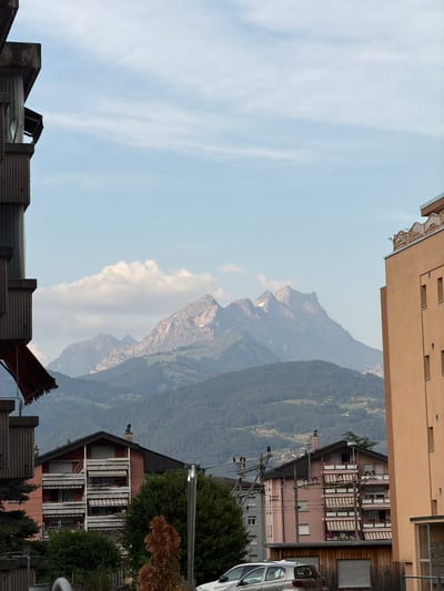 The image shows a view of mountains in Monthey, Switzerland, with buildings in the foreground. A large, grey, brutalist-style building is partially visible on the left. To the right, two residential buildings are prominent. The building on the left of this pair is pink with multiple balconies, each covered by striped awnings. The building on the right is a lighter, peach color with a decorative railing on its roof. The background is dominated by a mountain range, with the peaks showing some snow. The sky is partly cloudy with a light blue hue. Power lines and poles are visible between the buildings, suggesting proximity to infrastructure. A silver car is parked in front of the pink building. The overall scene depicts an urban environment with a natural mountainous backdrop.