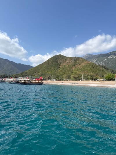 The image depicts a coastal scene in Kumluca, Turkey, featuring a beach bordered by a mountain range on one side and the Mediterranean Sea on the other.
The beach is light-colored and appears to be composed of sand or small pebbles. A few structures, possibly beach huts or buildings, are visible along the coastline.
The mountain range rises steeply behind the beach, covered in green vegetation. The sky is blue with scattered white clouds. The sea is calm with a deep blue color.
A long pier runs along the beach. It appears to accommodate several large boats or yachts. Red flags are visible on the vessels.