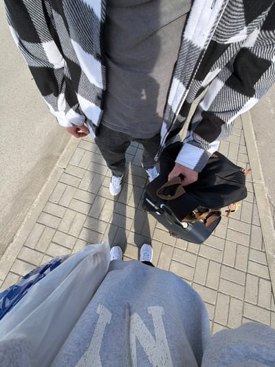 The image, taken from a high-angle perspective, shows two individuals standing on a brick-paved surface. On the left, a person is wearing a black and white plaid flannel shirt, a grey top, and dark trousers, holding a black bag on top of a suitcase. The suitcase is on wheels and has a retractable handle. They are wearing white Adidas sneakers. On the right, another person is wearing a grey hoodie and white sneakers. They are holding a large white plastic bag. The background is a brick-paved surface, partially with pavement and partially with the street. Given the absence of specific contextual details, the location cannot be definitively placed in Boldino, Russia.