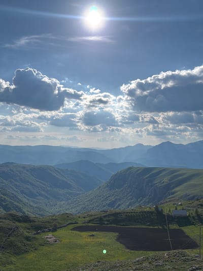 The image shows a landscape near Boričje, Montenegro. In the foreground, there are steep, green, mountainous hillsides with evidence of cultivated fields and a small building. In the mid-ground, there are layers of blue-grey mountains fading into the distance. The sky is a bright blue with puffy white clouds extending vertically, and the sun is visible with a lens flare effect.