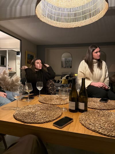 The image depicts an indoor social gathering, likely an evening dinner or drinks, in Saint-Étienne, France. Two women are seated around a wooden dining table in what appears to be a home or restaurant setting.
One woman, with long dark hair, wears a light-colored, ribbed turtleneck sweater and dark trousers. She is seen from the chest up, with her hands clasped in front of her, looking attentively towards the side. The second woman, also with long dark hair, wears a dark, fuzzy top. She is positioned closer to the viewer, looking towards the camera with a neutral expression, with her hands near her head, possibly adjusting her hair.
The wooden table is set with several items. Two empty wine bottles, one green and one lighter, stand prominently. At least four wine glasses are visible, some empty, and one possibly containing white wine. Four round placemats, made of woven natural fibers, are arranged on the table. Two smartphones are placed face down. A decorative element resembling fluffy pampas grass is also on the table.
In the background, a large, circular pendant light fixture made of woven material hangs from the ceiling, providing ambient lighting that suggests it is nighttime. An arched mirror on the wall reflects a patterned background, and a framed piece of art with a yellow border is visible further back. A leopard print cushion or bag rests on a seat next to one of the women.
The scene conveys a relaxed and convivial atmosphere. The text on the wine bottle labels is largely unreadable, though parts of "MOS" and "BLANC" are faintly visible on one.