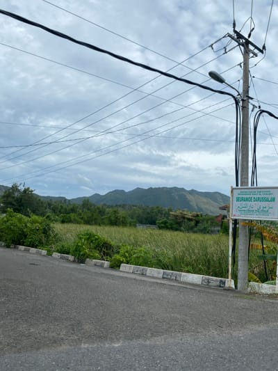 The image shows a rural scene in Beuradeun, Indonesia.  A utility pole with power lines dominates the left side of the image.  A sign, "SEURAMOE DARUSSALAM," with Arabic script, is mounted on a concrete post to the right of the pole. Vegetation is present along the roadside. In the background, a mountain range is visible under a cloudy sky.  A paved road extends into the foreground.