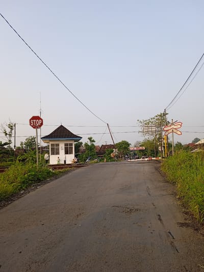The image depicts a rural road crossing a railroad track in Sidorejo, Indonesia. A small, white control booth with a black roof is located to the left of the road, adjacent to a stop sign. A railroad crossing sign stands to the right of the road. Vegetation lines both sides of the road. People are present near the control booth and on the railroad track. Several motorcycles are present beyond the gate. The road itself is asphalt, showing signs of wear and tear. Overhead power lines extend across the scene.
