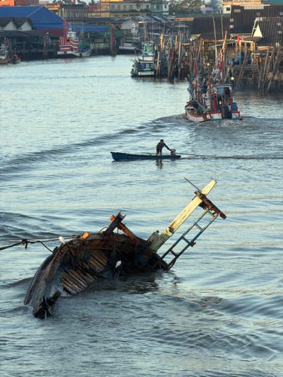 The image depicts a serene river scene in Pak Nam Chumphon, Thailand, at sunset. In the foreground, a partially submerged wooden boat, its hull tilted and listing to the side, rests in the water, with two white egrets perched on its debris. The weathered wood and exposed ribs of the boat suggest it has been damaged or is derelict.

Further out on the river, a small, dark blue boat with a single occupant is being propelled by an outboard motor. The person, dressed in dark clothing, is standing and appears to be steering the vessel.

In the background, a larger, red and white fishing vessel is seen navigating the water, leaving a wake behind it. This boat is equipped with numerous fishing gear and lights. Along the riverbanks, a dense collection of wooden stilt houses and docks, characteristic of a fishing village, lines the shore. Various other fishing boats are moored or docked amongst the structures. The warm light of the setting sun casts a soft glow over the entire scene, highlighting the tranquil yet industrious atmosphere of the location.