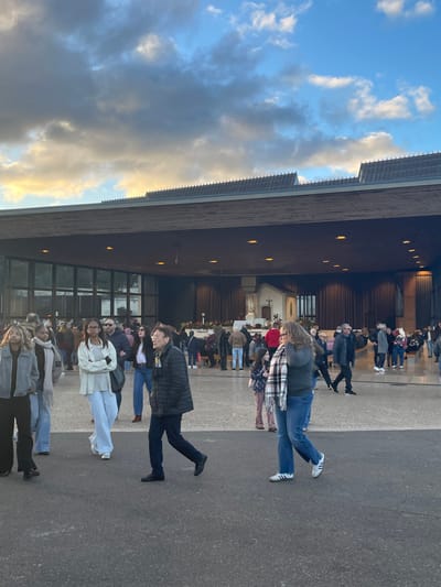 The image captures an outdoor area in front of the Basilica of the Holy Trinity at the Sanctuary of Fátima, Portugal, during what appears to be late afternoon or early evening. The sky above is partially cloudy, featuring patches of blue interspersed with grey and white clouds, some exhibiting a golden hue on their undersides.
The dominant structure is the modern basilica, visible in the background, characterized by a broad, dark-colored roof with a distinctive serrated or ridged edge, extending forward as a deep canopy over the main entrance. The building's facade includes large sections of dark-framed glass panels on the left, reflecting a muted environment. To the right, beneath the canopy, a recessed entrance area reveals a visible interior featuring a small, light-colored structure resembling a chapel or shrine with a statue (likely of the Virgin Mary), decorated with floral arrangements. Benches and individuals are discernible within this interior space.
Numerous people are present across the paved ground in front of the basilica, indicating a moderately crowded scene. Most individuals are in motion, walking in various directions. In the immediate foreground, from left to right:
1. A person with long, light-colored hair, wearing a grey jacket and dark pants, walks towards the right.
2. Next to them, a person with dark hair, wearing a light grey scarf and light-colored clothing, is partially visible.
3. A woman with dark hair, wearing a light-colored jacket over a lighter top and light blue wide-leg pants, walks towards the right.
4. A man wearing a dark puffer jacket, dark pants, and glasses, with a small yellow pin on his jacket, walks towards the right.
5. A woman with long, curly dark hair, wearing a dark top, blue jeans, and a black, white, and pink/red checkered scarf, walks towards the left. She wears white sneakers with black stripes. A child is partially obscured next to her.
Further back, more individuals are scattered across the paved area and gathered closer to the basilica entrance, some seated on benches within the building. The overall scene depicts a public gathering in a religious site.