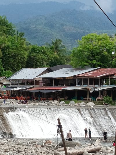 The image depicts a dynamic riverside environment in Meurah, Indonesia, characterized by a constructed waterfall, multiple open-air structures, and a mountainous backdrop. The foreground features a broad riverbed composed of numerous irregularly shaped rocks and pebbles. A wooden utility pole, secured with ropes, stands vertically on the left, with wires extending diagonally towards the upper right. Several individuals are present in the shallow water or on the rocky bank; these include a person in dark swimwear, two individuals in light and dark casual attire, and a person in a long dark dress with a head covering. Above the riverbed, a concrete weir spans the width of the frame, over which water cascades downwards, forming a wide, turbulent white waterfall.
Directly above the waterfall, a row of multi-story buildings, likely commercial establishments such as cafes or restaurants, lines the riverbank. These structures feature corrugated metal roofs, some appearing weathered silver-gray and others rusted reddish-brown. Their lower levels are open, revealing multiple sets of orange and light blue plastic tables and chairs. Numerous people are seated at these tables or standing on the rocky bank in front of the buildings, particularly on the left side of the frame. Behind these structures, dense, vibrant green foliage and various trees, including palm varieties, cover the landscape. The background consists of successive layers of distant, forested mountains, which appear hazy and blueish. A spherical white object, possibly a light fixture, is visible hanging in the upper right portion of the image, amidst the trees.