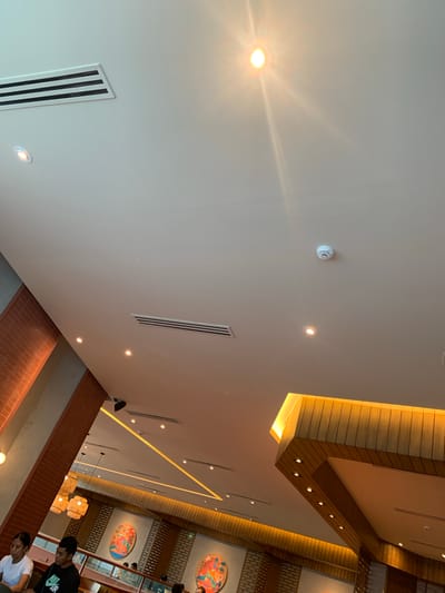 The image depicts the interior of a restaurant in Angeles City, Philippines. The perspective is from a low angle, looking up at the ceiling. The ceiling is white and features several recessed lights and air conditioning vents. A white circular smoke detector is also visible on the ceiling.
The restaurant has a modern design with warm lighting. On the right side of the image, a section of the ceiling is illuminated with a golden hue, highlighting a wooden slatted structure. In the background, decorative round wall art with colorful depictions is visible.
Two young people, a female and a male, are seated at a table in the foreground on the left. The female is wearing a white t-shirt, and the male is wearing a black t-shirt with a white logo. Other patrons are visible in the background, suggesting a busy dining environment. The overall ambiance appears to be casual and inviting.