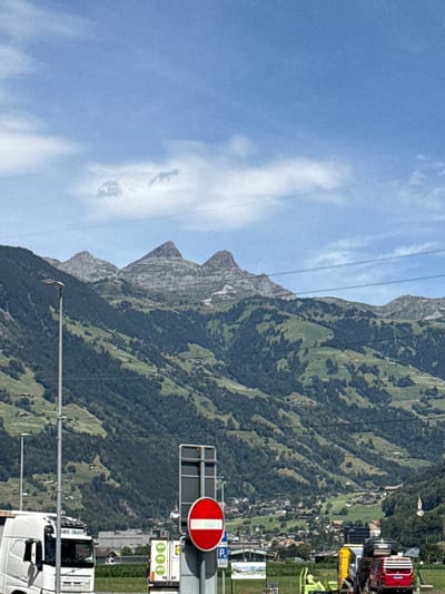 The image captures a scenic view of Schattdorf, Switzerland, with a focus on the transition between urban infrastructure and the natural mountain landscape. In the foreground, a white semi-truck, branded with "ORIS TRANS", is parked on the left side of the frame. To the right, a red and white circular traffic sign indicates "no entry" or "do not enter". Behind the truck, partially visible, is a white trailer with green markings displaying percentages. Further to the right, a yellow vehicle is loaded with cars, one of which is red, and a green tractor is visible.

The middle ground presents a village nestled at the base of a steep, verdant mountain range. Numerous houses with red roofs are scattered across the slopes, interspersed with dark green pine forests. A church spire with a white steeple is visible in the village.

The background is dominated by majestic, rugged mountains, characterized by rocky peaks and lush green slopes. The sky is a clear blue, with a few wispy white clouds. Electrical power lines are visible stretching across the frame, indicating the presence of infrastructure within this picturesque setting. The overall scene suggests a sunny day in a mountainous region of Switzerland.