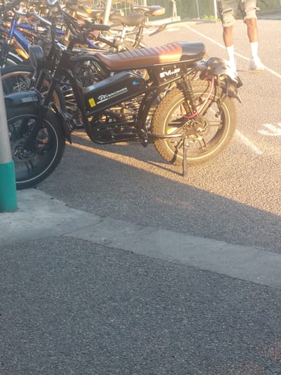 The image shows a bicycle rack filled with various bicycles, including an e-bike. The e-bike is a black frame model with a tan seat and a battery pack labeled "EVERCROSS." The bicycles are parked on an asphalt surface next to a concrete curb, suggesting a street or parking area. A portion of two people's legs can be seen in the upper left corner, wearing white socks. The light suggests it's either early morning or late afternoon. The location is likely Altdorf, Germany, based on the "ACHSELRAD" sticker on the bicycle, which could be a shop in that area.