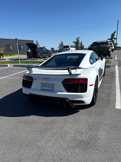 The image, which appears to be rotated 90 degrees clockwise from its original portrait orientation, depicts a bright, sunny day in a paved parking area. When viewed correctly (sky at the top), the central subject is a white Audi R8 sports car, captured from its rear passenger side, parked at an angle. The car features a prominent black rear spoiler, black multi-spoke wheels, and distinctive black mesh grilles on the rear fascia, complemented by black exhaust tips and diffuser elements. Its license plate, bearing the text "A99 9LD" and "Beautiful British Columbia" with a Canadian flag, clearly indicates the vehicle's registration in British Columbia. The Audi rings logo and "R8" emblem are also visible on the trunk lid.
In the middle ground, several other vehicles are parked. A dark-colored SUV is notable, with its trunk open, and a person wearing a striped shirt is seen interacting with its contents. Further behind, parts of a white vehicle and a motorcycle can be discerned. To the left, a modern building with dark grey siding and rectangular windows stands, adjacent to which a "No Parking" sign (red circle with a 'P' and a diagonal line) is visible on a pole. Scattered evergreen trees and a tall black lamppost contribute to the background.
The expansive blue sky, coupled with the strong sunlight and clear, dark shadows cast by the vehicles, suggests a mid-day or early afternoon setting. The pavement is dark asphalt with white parking lines. Another individual is partially visible near the lower-left corner of the frame. The scene captures a typical moment in a parking lot, consistent with the described location of West Vancouver, Canada.